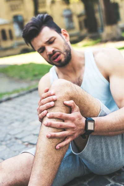 Handsome sportsman suffering from pain and touching injured leg while sitting on pavement — Stock Photo