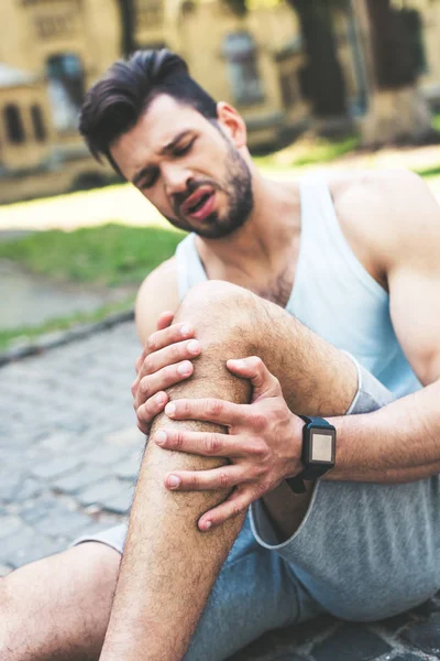 Beau sportif souffrant de douleur et touchant jambe blessée tout en étant assis sur la chaussée — Photo de stock