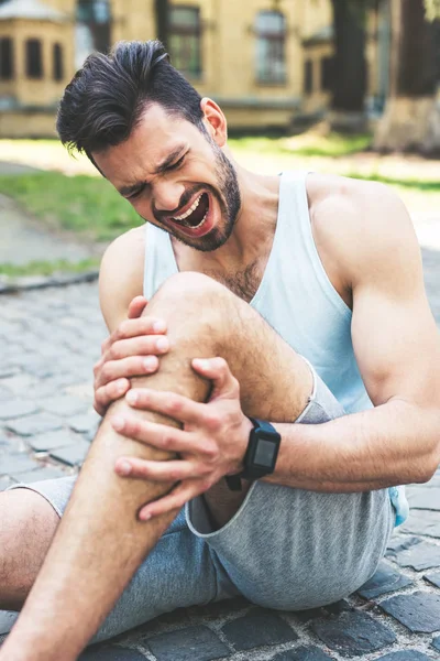 Young sportsman suffering from pain and screaming while sitting on pavement and touching injured leg — Stock Photo