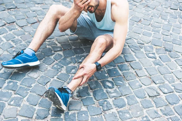 Joven deportista que sufre de dolor mientras está sentado en la pasarela, tocando la pierna lesionada y sosteniendo la mano en la cara - foto de stock