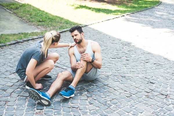 Woman in sportswear touching shoulder of injured sportsman sitting on pavement and suffering from pain — Stock Photo