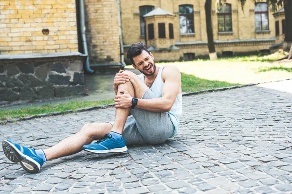 Beau sportif criant tout en étant assis sur la chaussée et touchant jambe blessée — Photo de stock