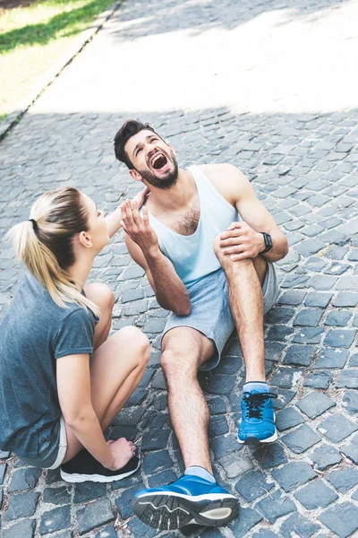 Young woman in sportswear calming sportsman screaming while sitting on pavement and touching injured leg — Stock Photo