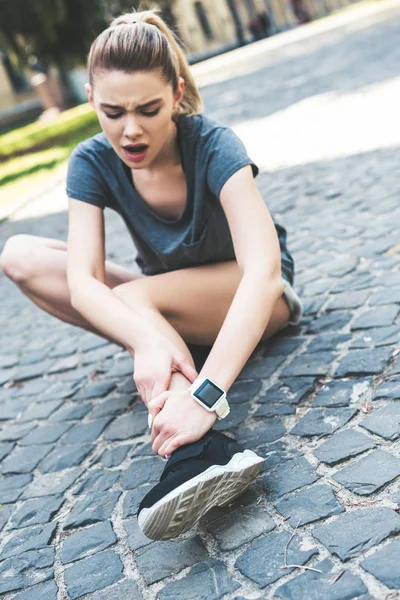 Young sportswoman suffering from pain while sitting on pavement and touching injured leg — Stock Photo