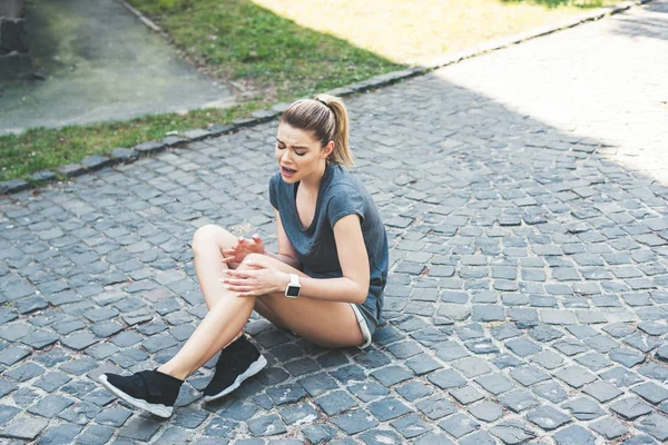 Young sportswoman sitting on pavement and suffering from pain while touching injured leg — Stock Photo