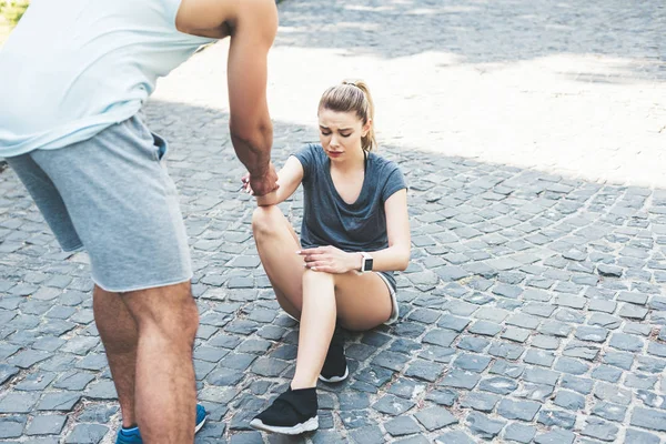 Coup de feu recadré de l'homme en vêtements de sport donnant la main à une sportive blessée assise sur le trottoir et souffrant de douleur — Photo de stock