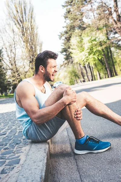 Young sportsman suffering from pain while sitting on pavement border and touching injured leg — Stock Photo