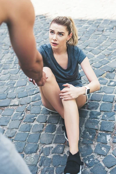 Cropped view of man giving hand to injured sportswoman sitting on pavement — Stock Photo