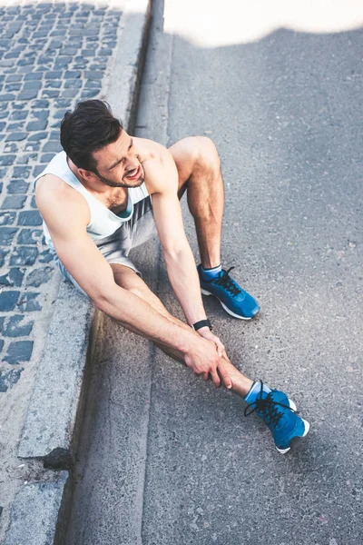 Вид зверху спортсмена, який страждає від болю, сидячи на кордоні і торкаючись травмованої ноги — стокове фото