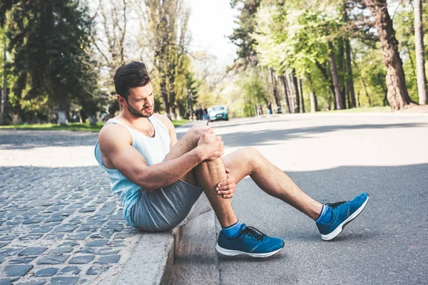 Young sportsman sitting on pavement border and touching injured leg — Stock Photo