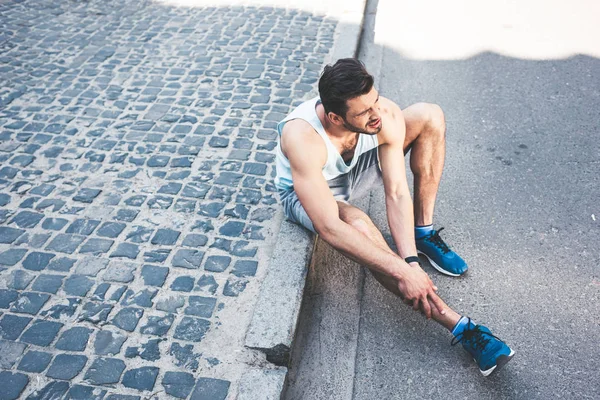 Deportista lesionado que sufre de dolor mientras está sentado en la frontera y tocando la pierna lesionada - foto de stock