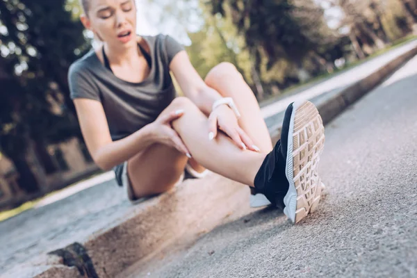 Foco seletivo da jovem esportista sentada na fronteira e tocando a perna ferida — Fotografia de Stock