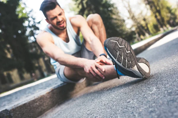 Selective focus of handsome sportsman sitting on border and touching injured leg — Stock Photo