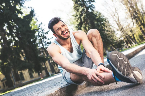 Deportista gritando sufriendo de dolor mientras está sentado en la frontera y tocando la pierna lesionada - foto de stock