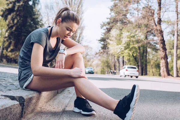 KYIV, UKRAINE - APRIL 25, 2019: Young sportswoman suffering from pain while sitting on pavement and touching injured leg. — Stock Photo