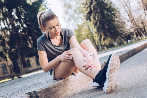 Грустная спортсменка, страдающая от боли, сидя на тротуаре и касаясь поврежденной ноги — стоковое фото