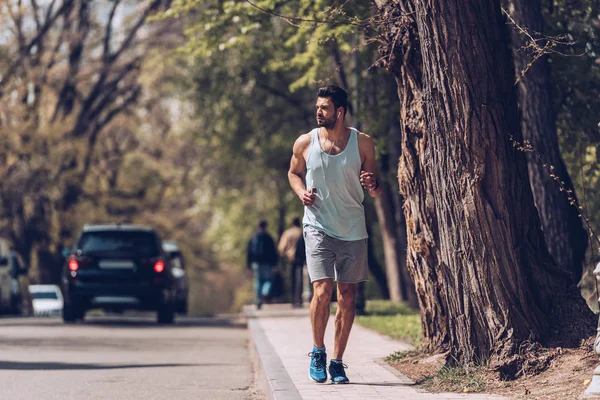 KYIV, UCRANIA - 25 DE ABRIL DE 2019: Guapo deportista corriendo por el pavimento cerca de la carretera y escuchando música en los auriculares . - foto de stock