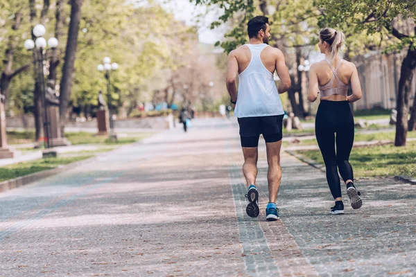 Вид сзади на мужчину и женщину в спортивной одежде, бегущих вдоль аллеи парка — стоковое фото