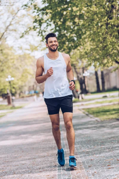 Desportista alegre sorrindo enquanto corre ao longo de passarela larga no parque — Fotografia de Stock