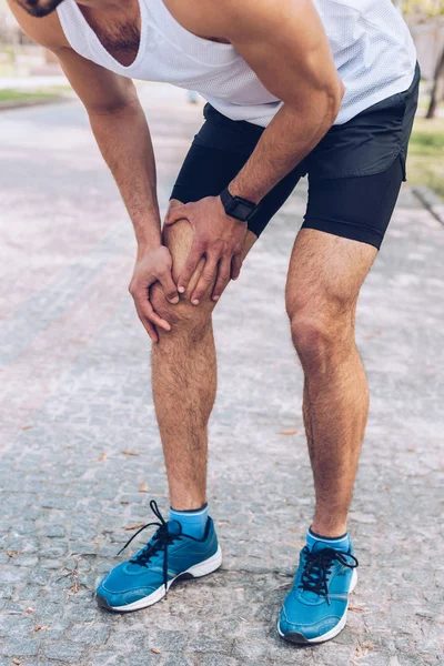 Частковий вигляд людини в спортивному одязі та кросівках, що торкаються травмованого коліна — стокове фото