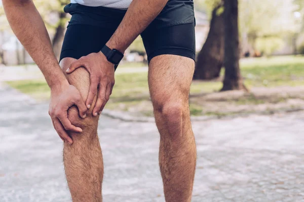 Cropped shot of young sportsman standing in park and touching injured knee — Stock Photo