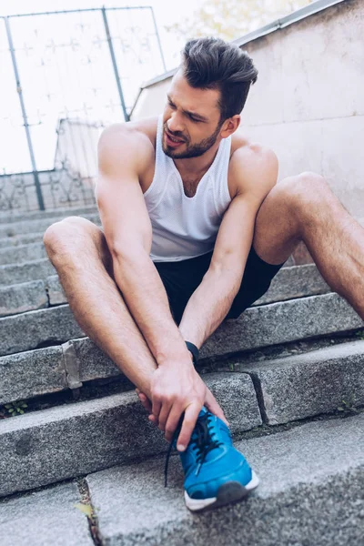 Upset sportsman sitting on stairs and suffering from pain while touching injured knee — Stock Photo