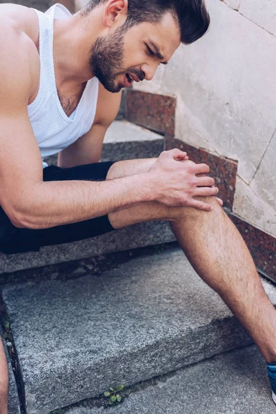 Молодой спортсмен трогает травмированное колено, сидя на лестнице и страдая от боли — стоковое фото