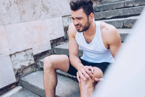 Selective focus of handsome sportsman suffering from hurt while sitting on stairs and touching injured knee — Stock Photo