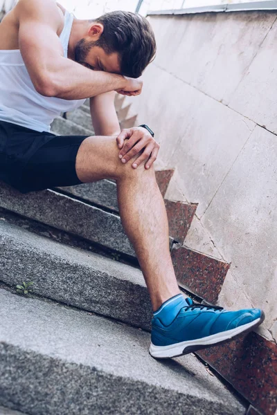 Травмований спортсмен, який страждає від болю, сидячи на сходах і тримаючи руку біля обличчя — стокове фото