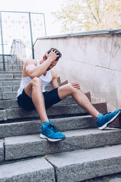 Deportista lesionado sentado en las escaleras y que sufre de dolor mientras sostiene la mano en la cabeza - foto de stock