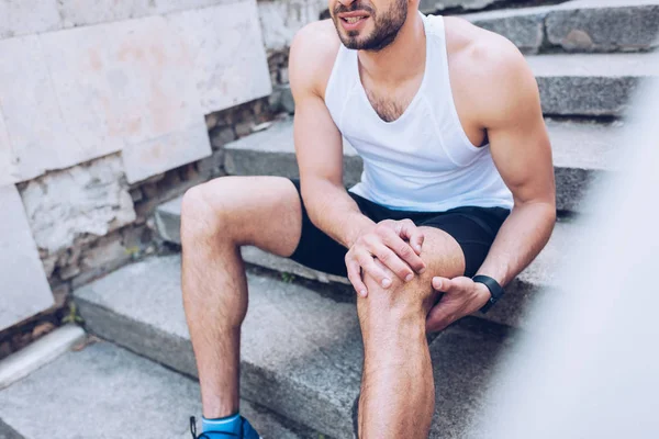 Частичный взгляд спортсмена, сидящего на лестнице и страдающего от боли при прикосновении к травме колена — стоковое фото