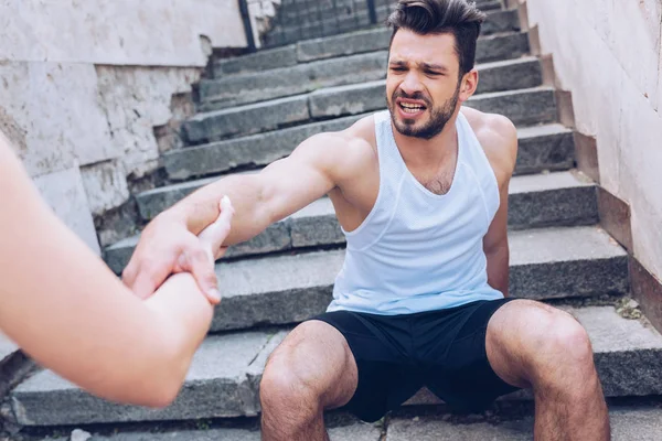 Partial view of woman giving hand to injured sportsman suffering from pain while sitting on stairs — Stock Photo