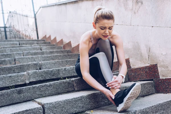 Attractive sportswoman sitting on stairs and suffering from pain while touching injured leg — Stock Photo
