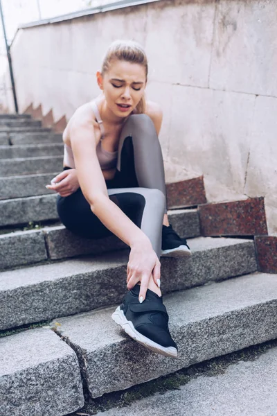 Injured sportswoman sitting on stairs and suffering from pain while touching leg — Stock Photo