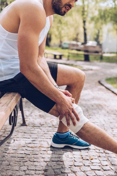 Plan recadré d'un sportif assis sur un banc et touchant un bandage élastique sur une jambe blessée — Photo de stock