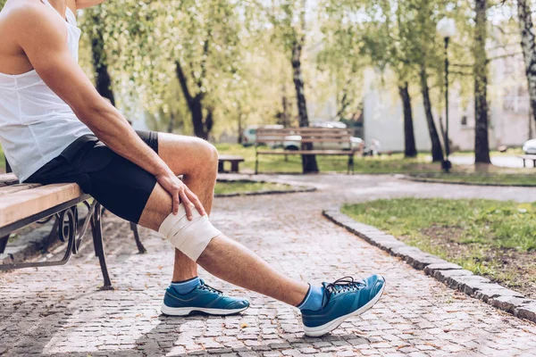 Partial view of injured sportsman sitting on bench and touching elastic bandage on knee — Stock Photo