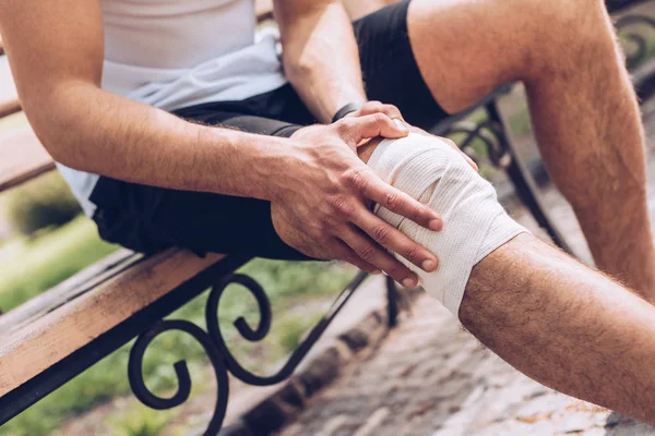 Cropped shot of sportsman sitting on bench and touching elastic bandage on injured knee — Stock Photo