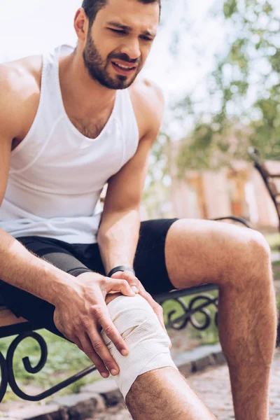 Расстроенный спортсмен, сидящий на скамейке и трогающий упругую повязку на травмированном колене — стоковое фото