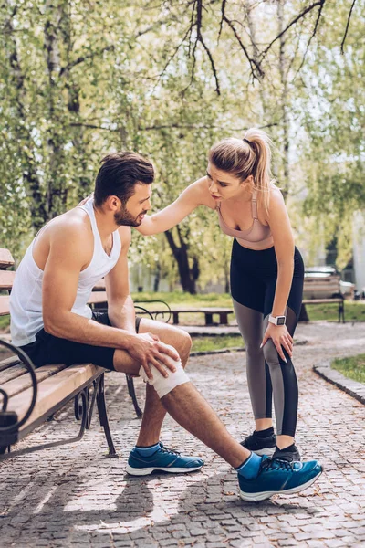 Bella donna in abbigliamento sportivo in piedi vicino a sportivo ferito seduto sulla panchina nel parco — Foto stock