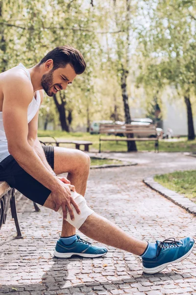 Young sportsman suffering from pain while sitting on bench and touching elastic bandage on injured knee — Stock Photo