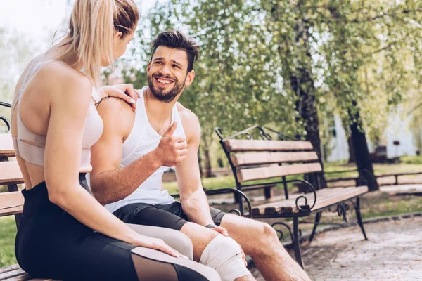 Verletzter Sportler lächelt und zeigt Daumen nach oben, während er auf Bank neben junger Sportlerin sitzt — Stockfoto