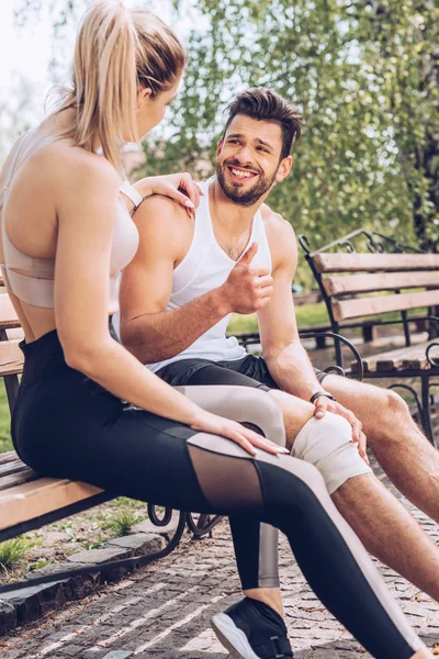Sportivo ferito sorridente e mostrando pollice in su mentre si siede sulla panchina vicino alla sportiva — Foto stock