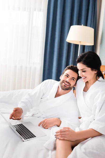 Hombre alegre gesto cerca de la computadora portátil y mirando a la mujer morena feliz en la habitación de hotel - foto de stock