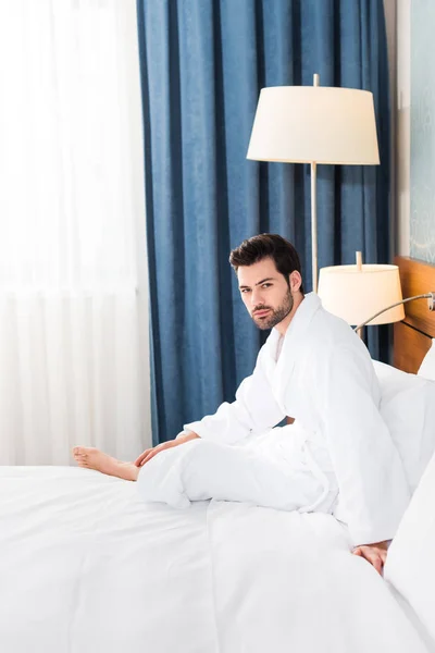 Hombre barbudo guapo mirando a la cámara mientras está sentado en la cama en la habitación de hotel - foto de stock