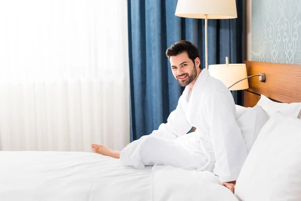 Happy bearded man looking at camera while sitting on bed in hotel room — Stock Photo