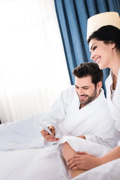 Hombre barbudo alegre usando teléfono inteligente cerca de mujer morena feliz - foto de stock