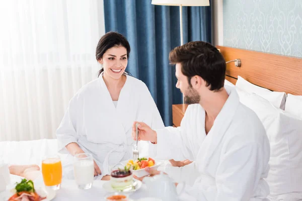 Selective focus of happy woman looking at bearded man holding fork near fruit salad — Stock Photo