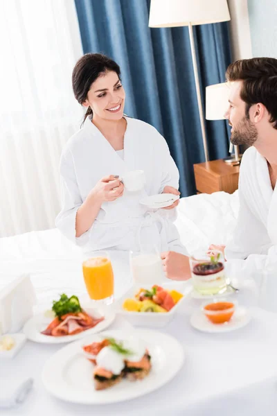 Mujer alegre sosteniendo platillo y taza mientras mira al hombre - foto de stock