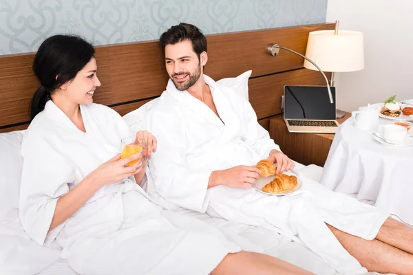 Cheerful man holding croissants while looking at woman with glass of orange juice — Stock Photo