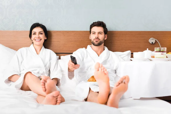 Enfoque selectivo del hombre alegre que sostiene el mando a distancia cerca de la mujer atractiva en la habitación del hotel - foto de stock